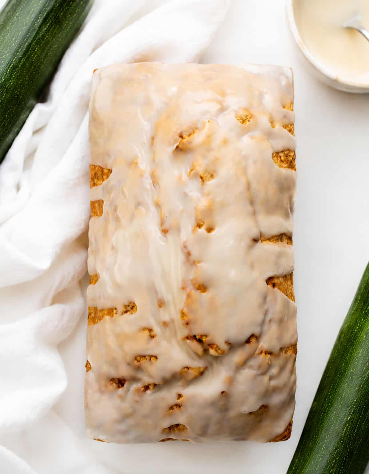 Whole Zucchini Pound Cake covered in glaze on a white table with zucchini from overhead.