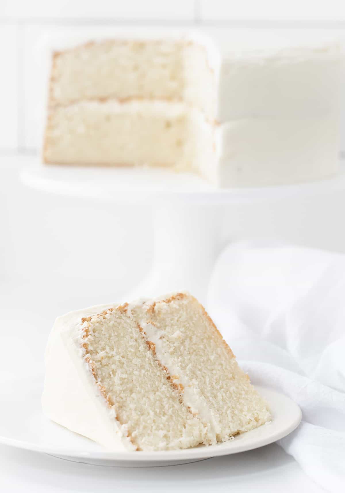 Piece of White Cake in Front of a Cut Into Cake on a Cake Stand. 