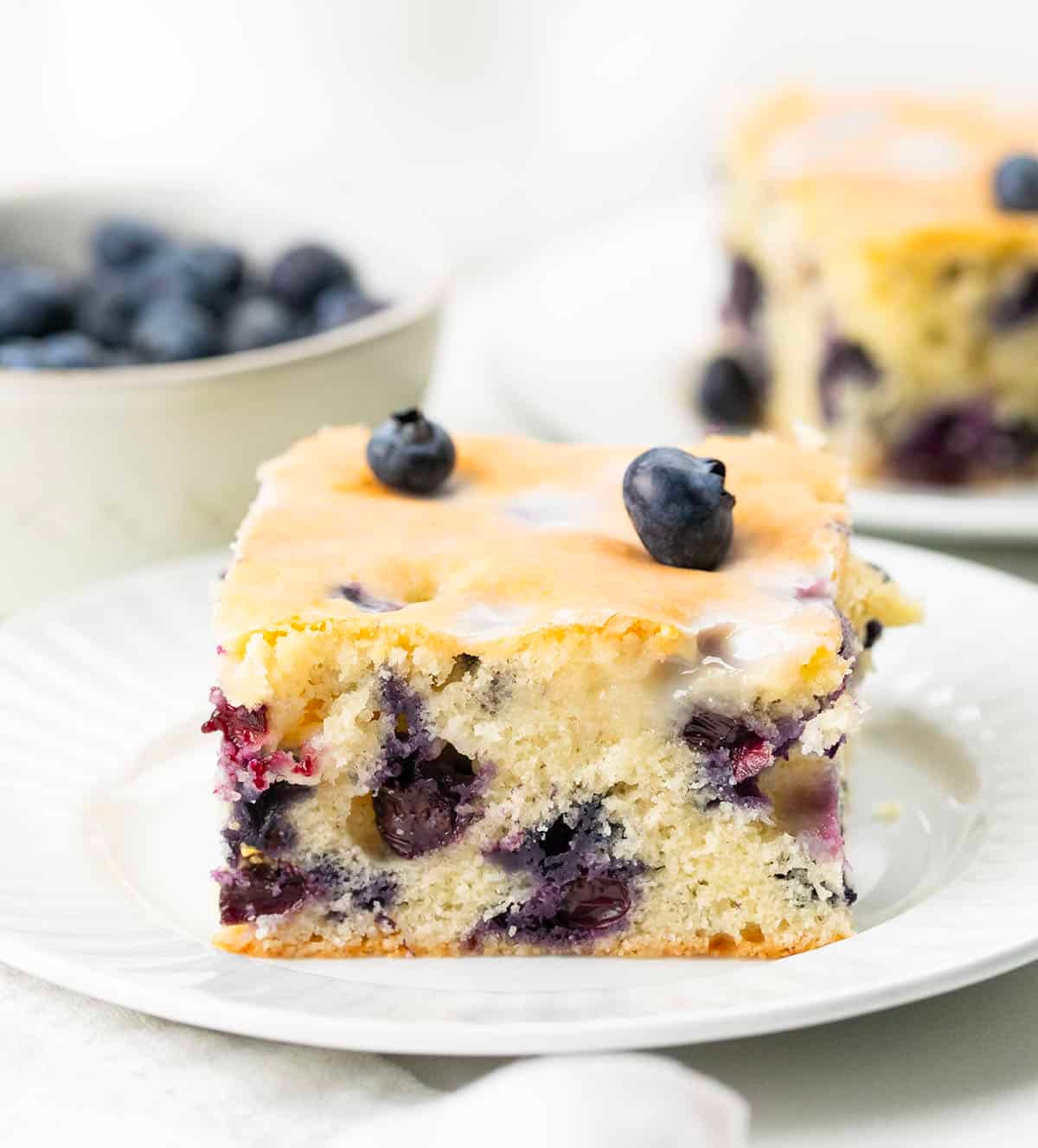 This Blueberry Breakfast Cake is tender, moist, and bursting with sweet, juicy blueberries in every bite! Slices of Blueberry Breakfast Cake on a white plates with fresh blueberries.
