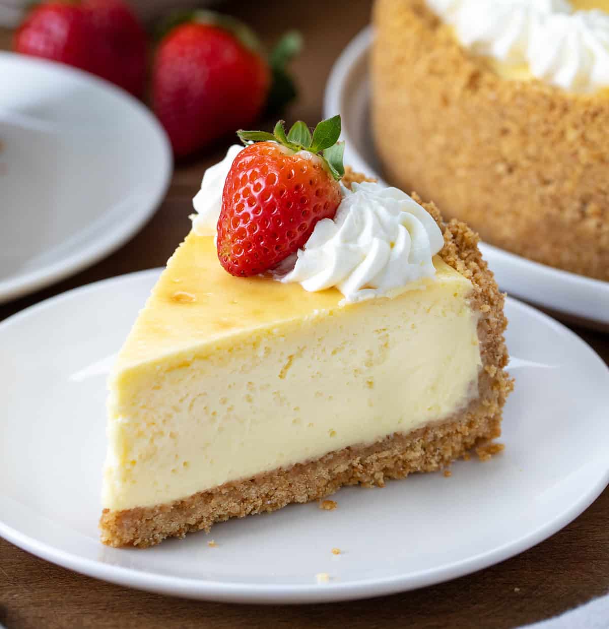 This Classic Cheesecake with Double Crust is perfect for anyone who loves a thick, buttery graham cracker base with a rich, creamy filling! Piece of Classic Cheesecake With Double Crust on a white plate on a wooden table.