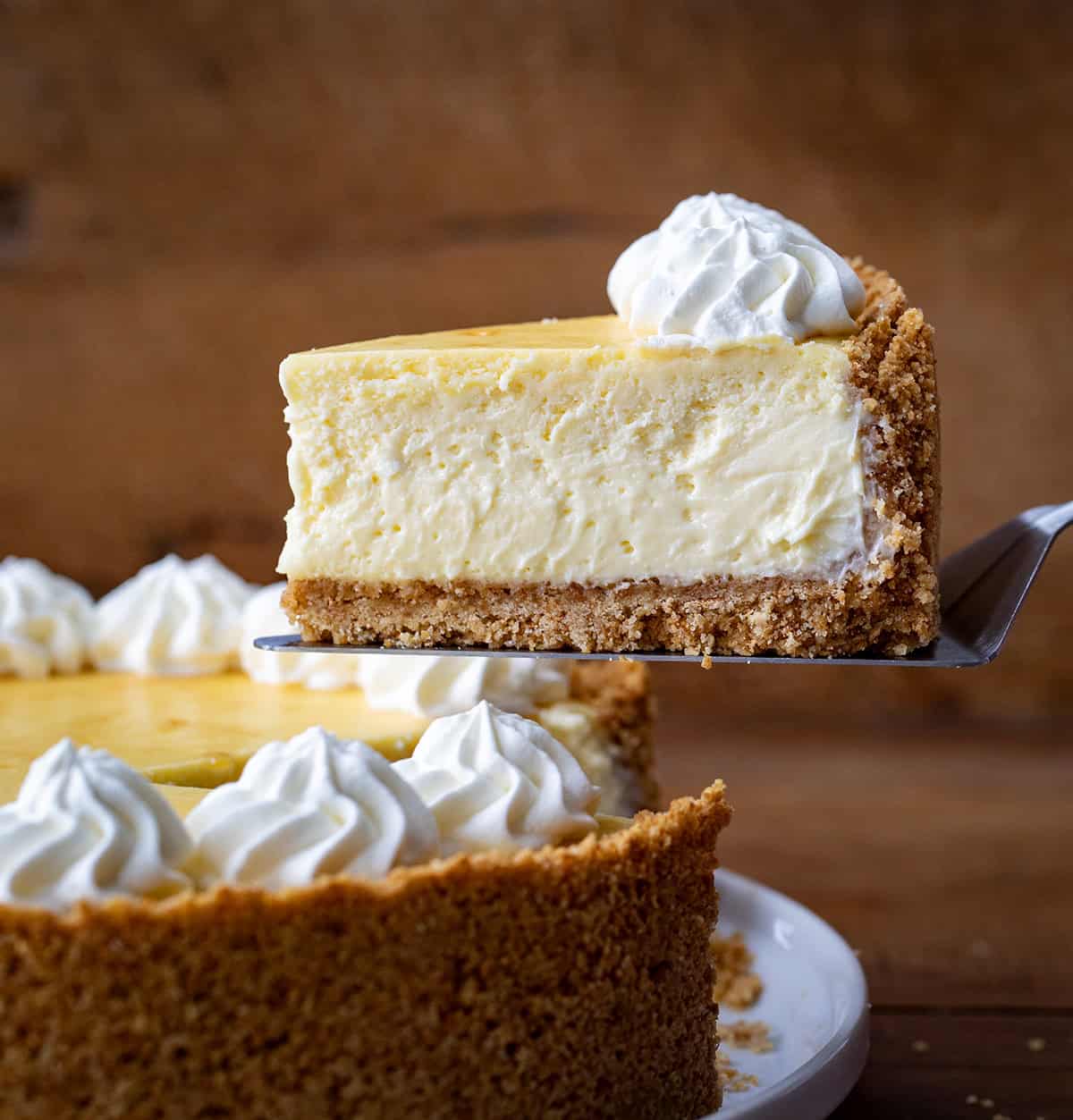 This Classic Cheesecake with Double Crust is perfect for anyone who loves a thick, buttery graham cracker base with a rich, creamy filling! Cake server picking up a piece of Classic Cheesecake With Double Crust from the serving plate.