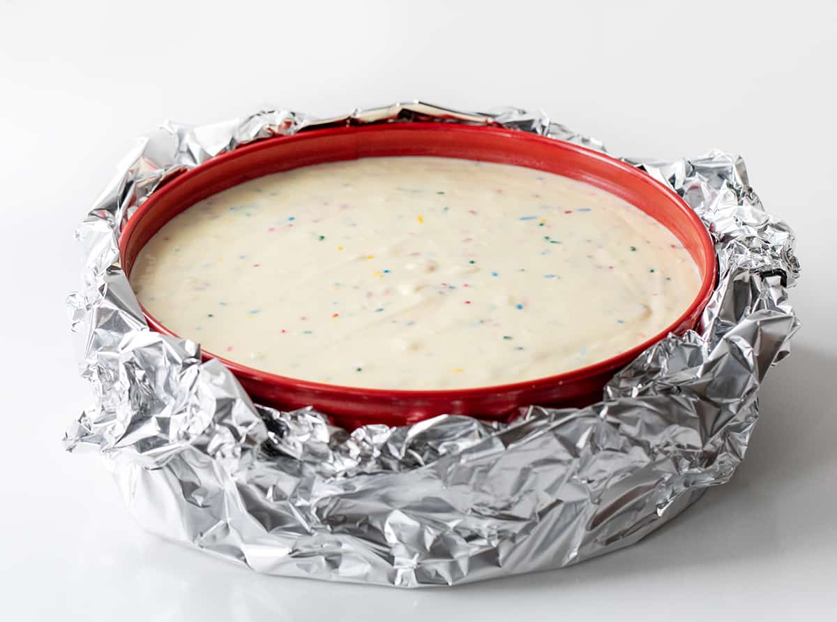 Raw Birthday Cheesecake Batter in a springform Pan with Foil around it on a White Counter.