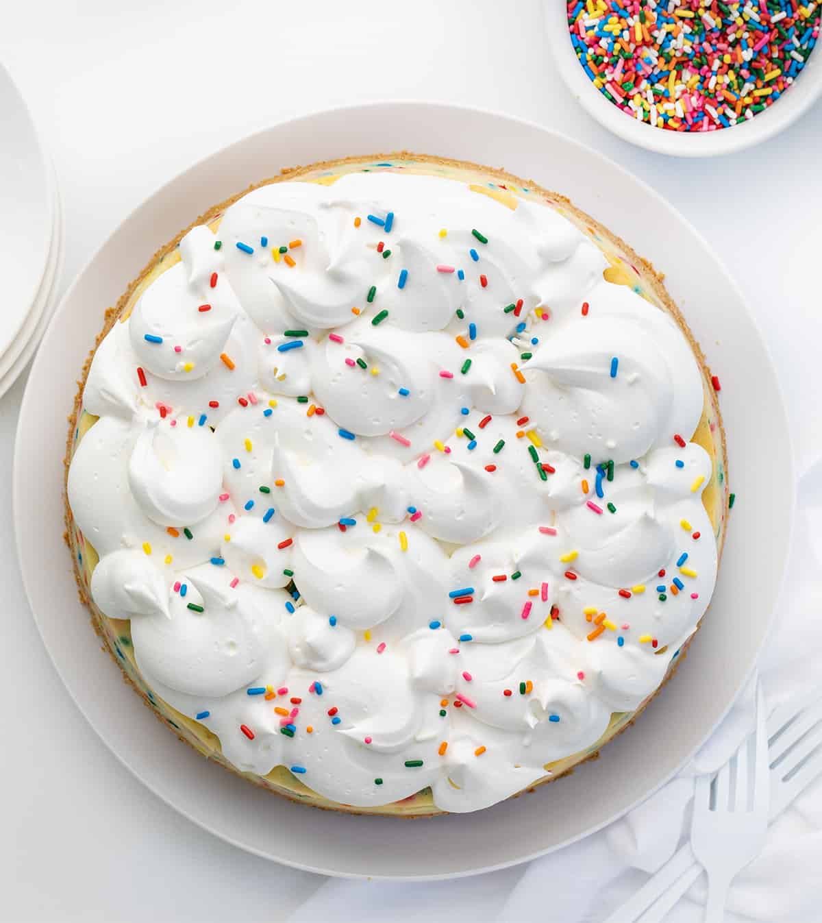 Overhead image of a Birthday Cheesecake Showing the Whipped Topping Dollops Covered in Sprinkles on a White Counter.