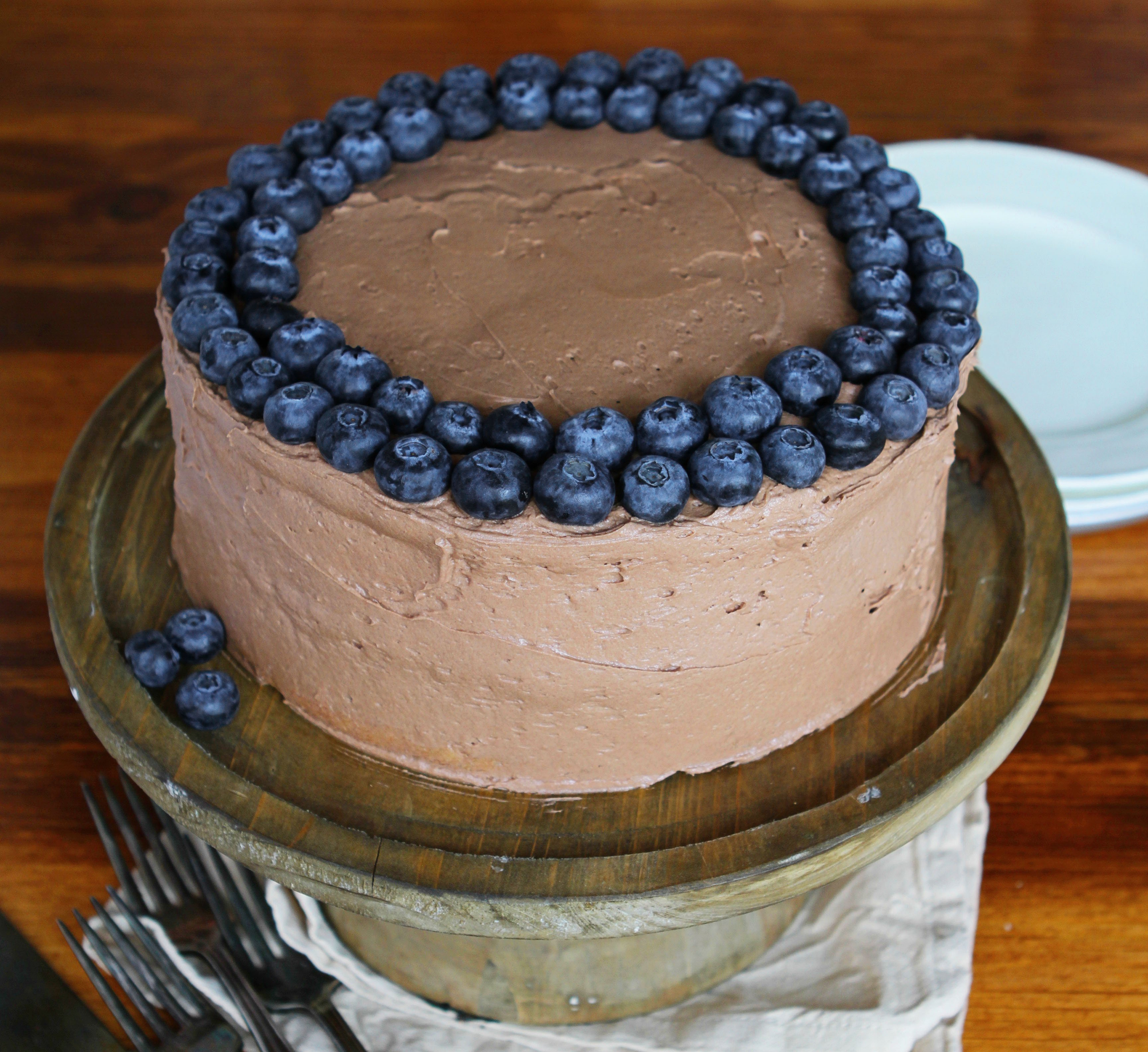 White Cake with Zucchini with Chocolate Frosting and Blueberries on Wooden Table with Plates in Background