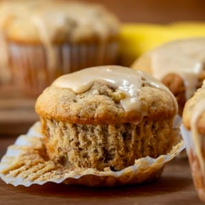 Half a peeled wrapper on a Banana Muffin With Vanilla Caramel Glaze on a wooden table surrounded by more muffins.