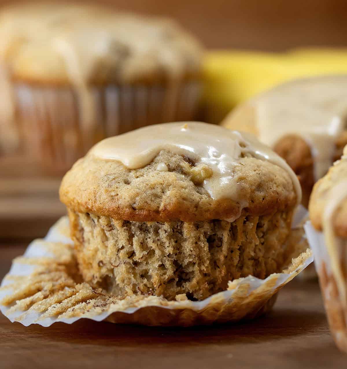 Half a peeled wrapper on a Banana Muffin With Vanilla Caramel Glaze on a wooden table surrounded by more muffins.