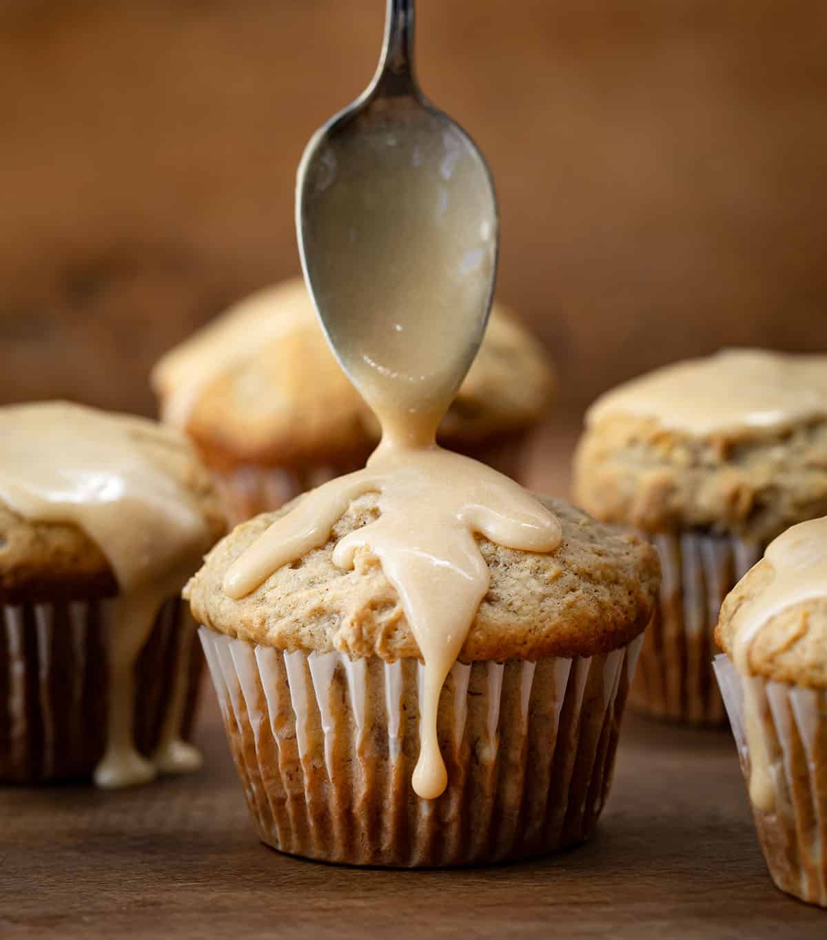 Drizzling vanilla caramel glaze over a banana muffin on a wooden table.