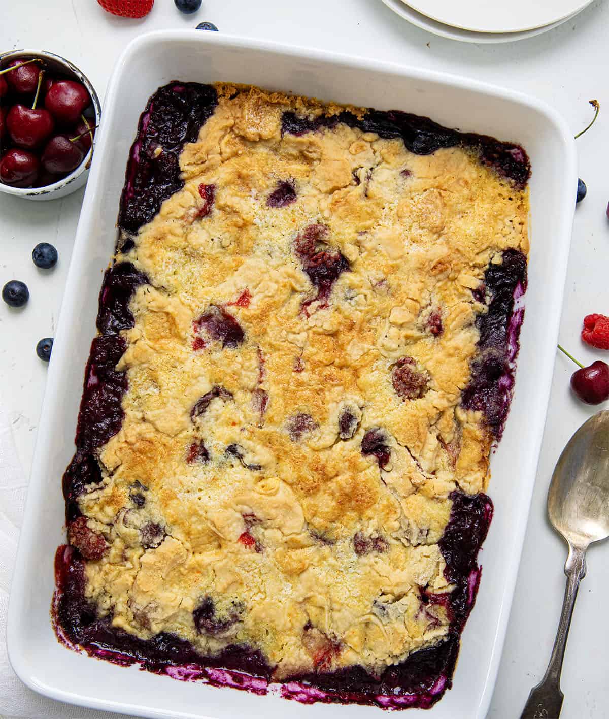 Pan of fruit cobbler on a white table with cherries and raspberries and blueberry from overhead. 