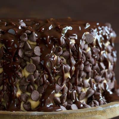 Brownie layer cake covered in cookie dough frosting with chocolate syrup drizzled over top on a rustic wooden cake stand.