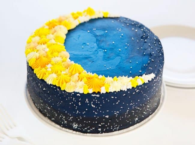 The side view of a dark blue galaxy cake with a moon piped on top!