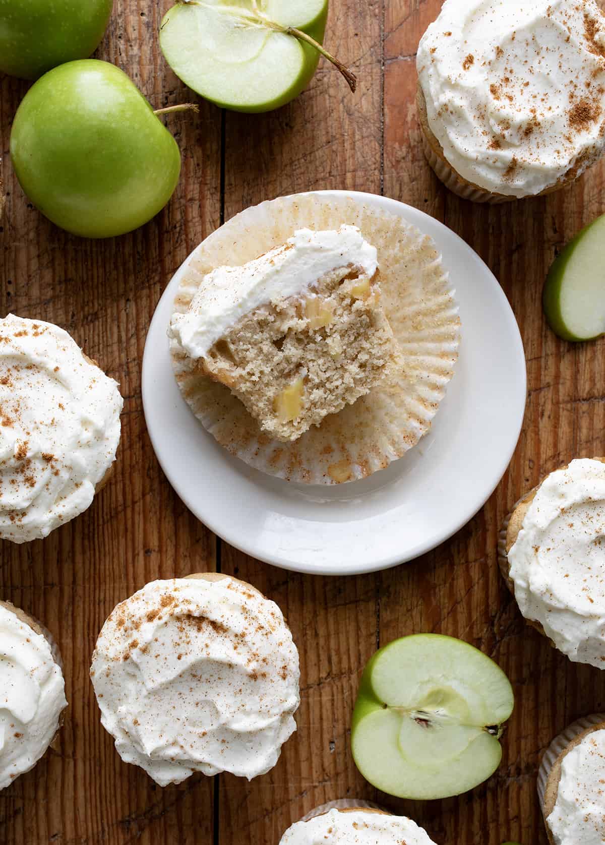 Apple Pie Cupcakes are perfectly spiced, tender cupcakes baked with apple pie filling and topped with stabilized whipped cream. So tender and delicious! They are reminiscent of a slice of apple pie but in convenient cupcake form! Apple Pie Cupcake Cut in Half on a Plate Surrounded by More Apple Pie Cupcakes and apples from Overhead.