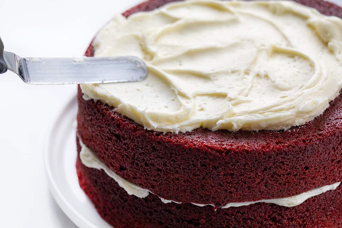 Small offset spatula smoothing cream cheese frosting over layered red velvet cake.