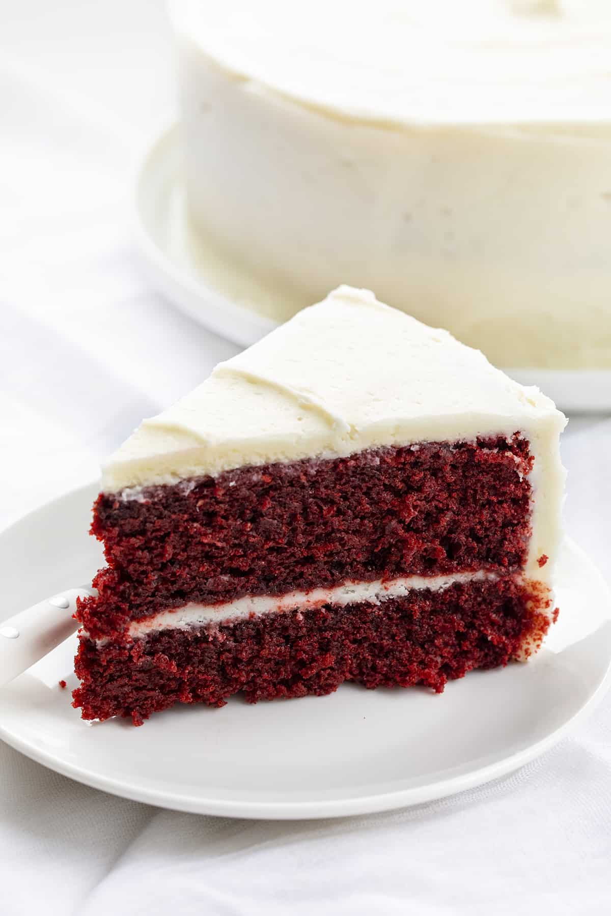Piece of Red velvet cake on a plate with the whole cake behind it in the background.