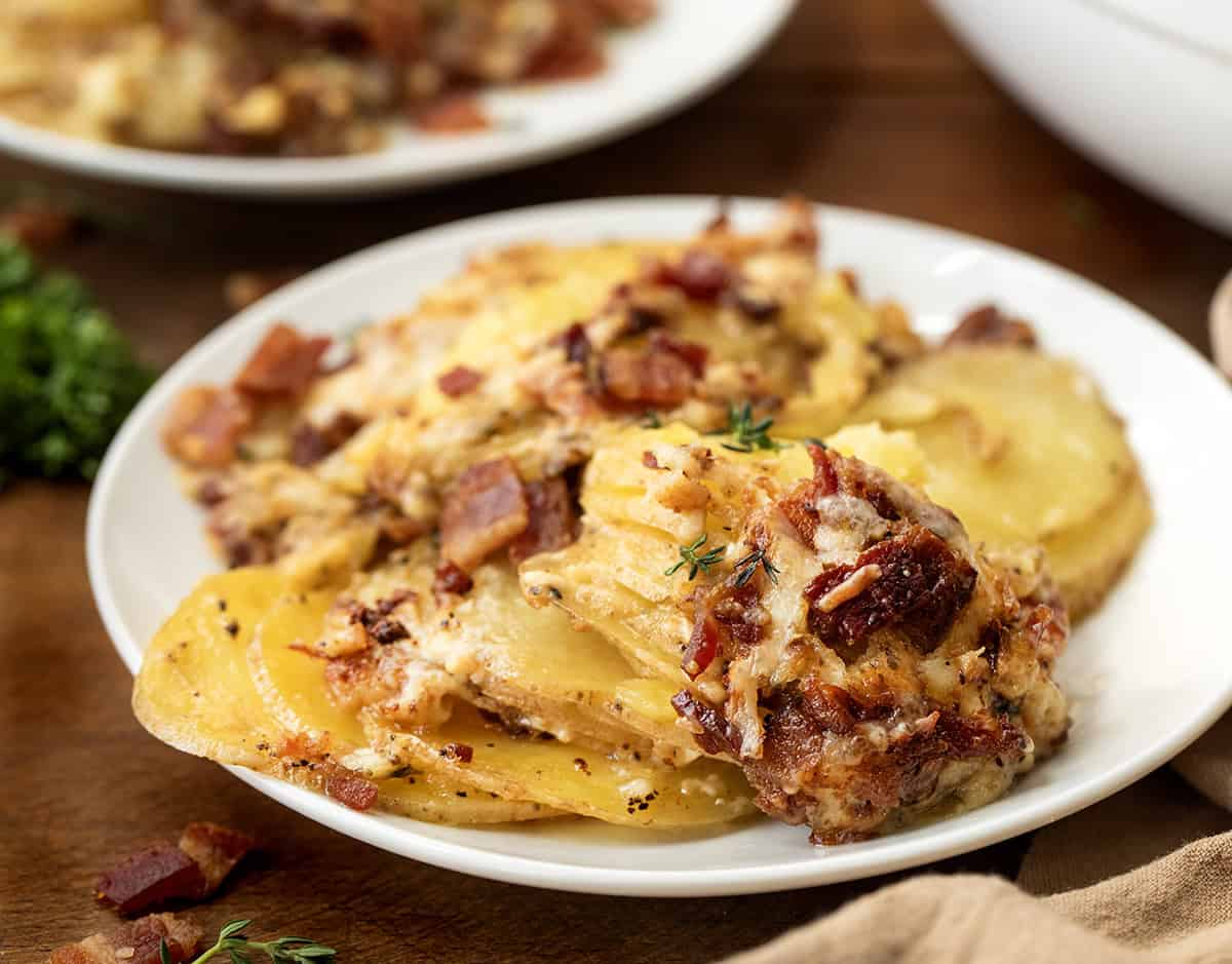 These Gruyere Scalloped Potatoes are a definite crowd pleaser! Loads of glorious gruyere cheese melting into thinly sliced Yukon gold potatoes and covered in bacon and thyme! White plate with Gruyere Scalloped Potatoes on it on a wooden table.