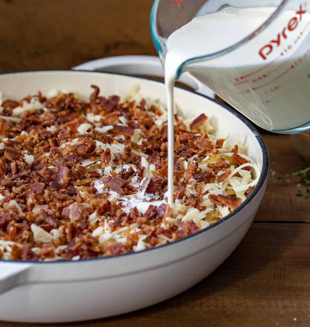 These Gruyere Scalloped Potatoes are a definite crowd pleaser! Loads of glorious gruyere cheese melting into thinly sliced Yukon gold potatoes and covered in bacon and thyme! Pouring cream into Gruyere Scalloped pan with potatoes and bacon.
