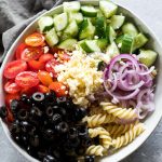 All the ingredients for the greek pasta salad in a large white bowl with grey napkin on the side