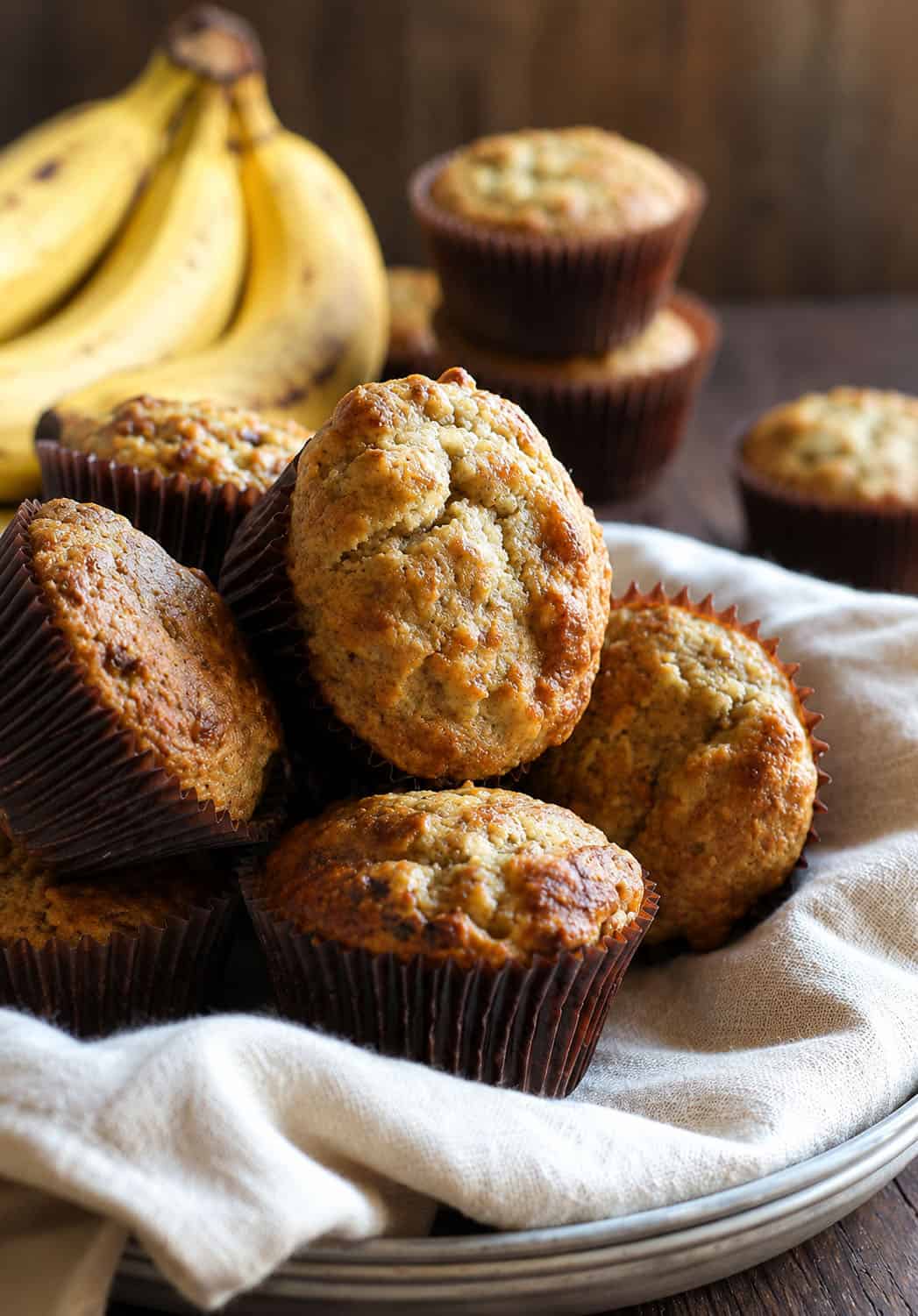 The purest, organic, delicious flavor of banana makes this the BEST Banana Muffin I have ever had! The secret to this recipe is DOUBLE the normal amount of banana! The Best Banana Muffins on a wooden table stacked with bananas in the background.