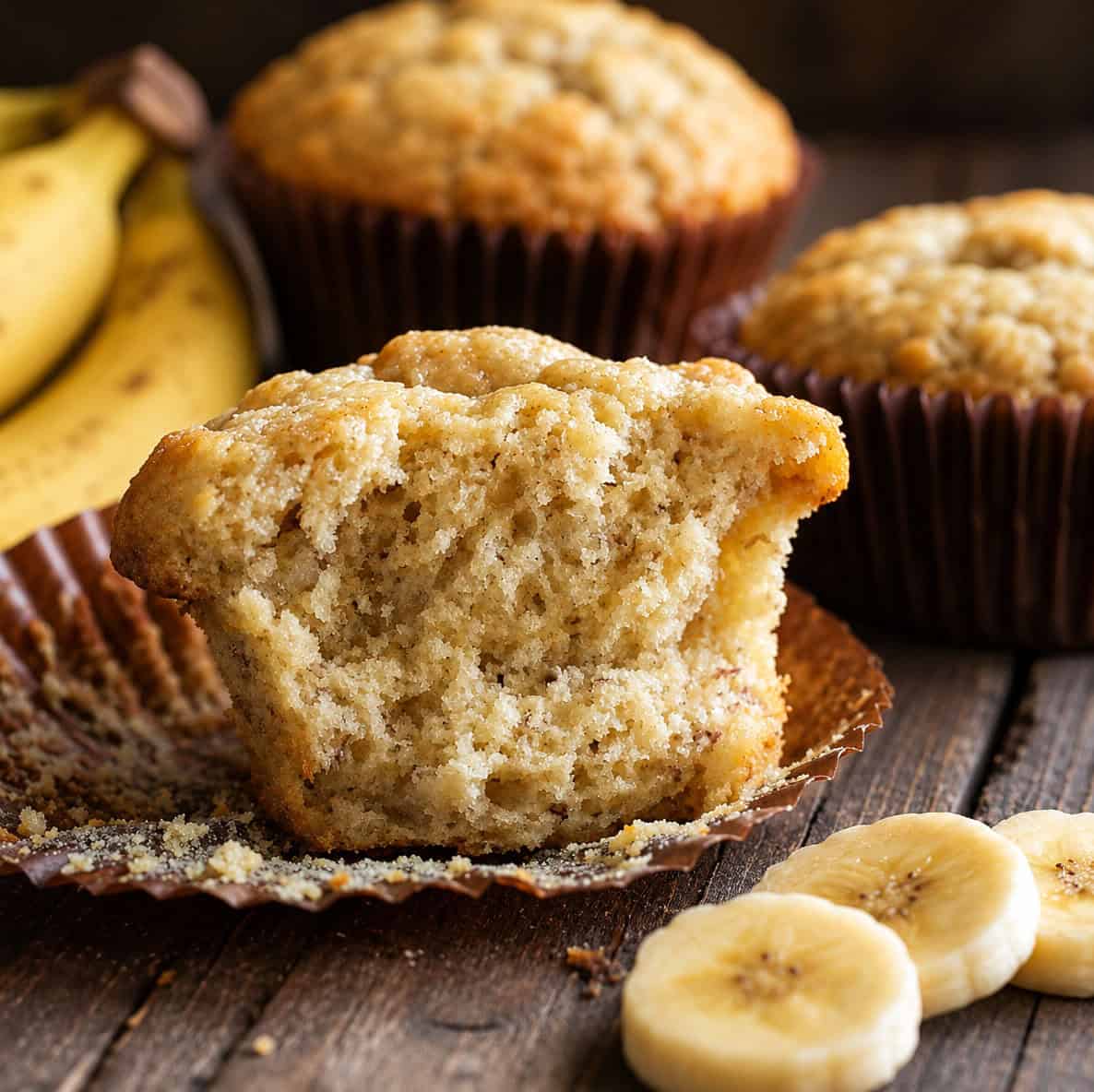 The Best Banana Muffins on a table with the main muffin halved showing inside texture.
