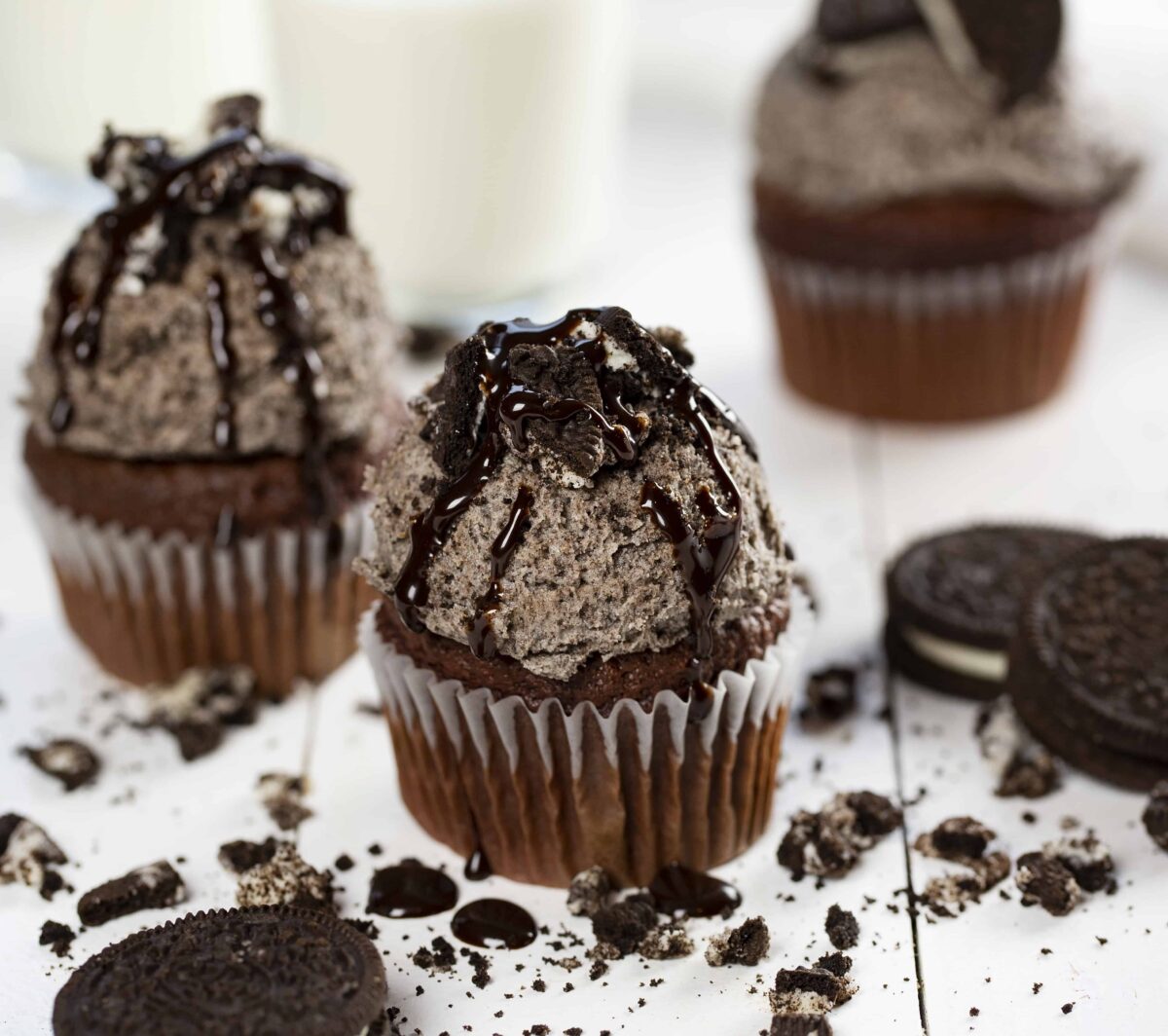 Oreo frosting covering chocolate cupcakes on a white table with milk and crushed Oreo's.
