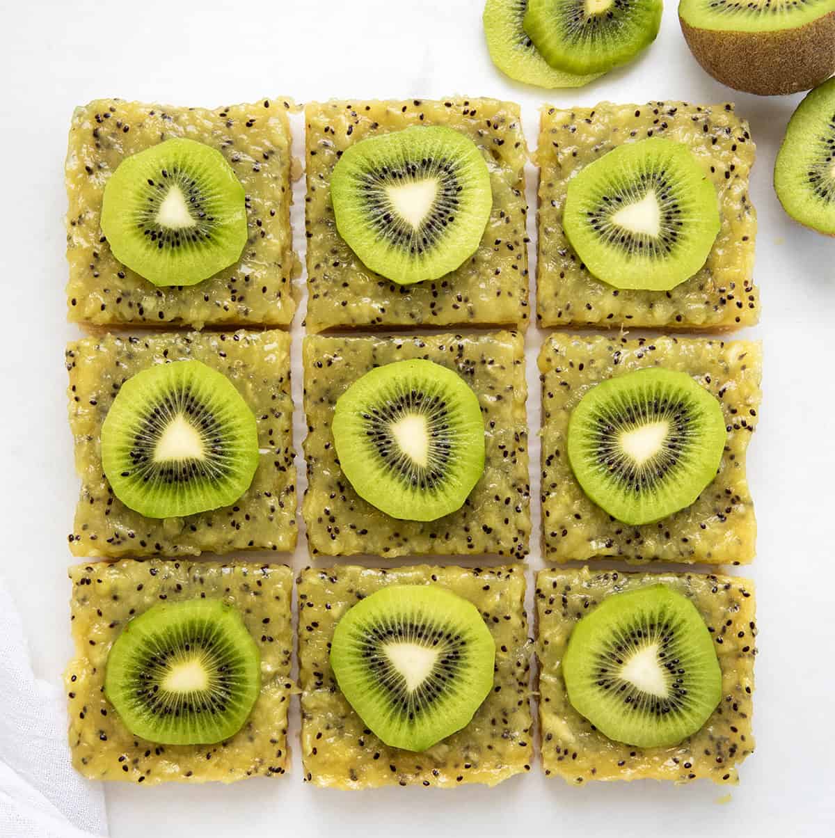 This Kiwi Bars recipe starts with a buttery shortbread crust topped with a smooth, sweet-tart kiwi that sets up perfectly. I like to serve the kiwi bars straight from the refrigerator, nice and chilled. The cool, creamy texture and bright flavor are extra refreshing that way! Kiwi Bars cut into squares on a white table from overhead.