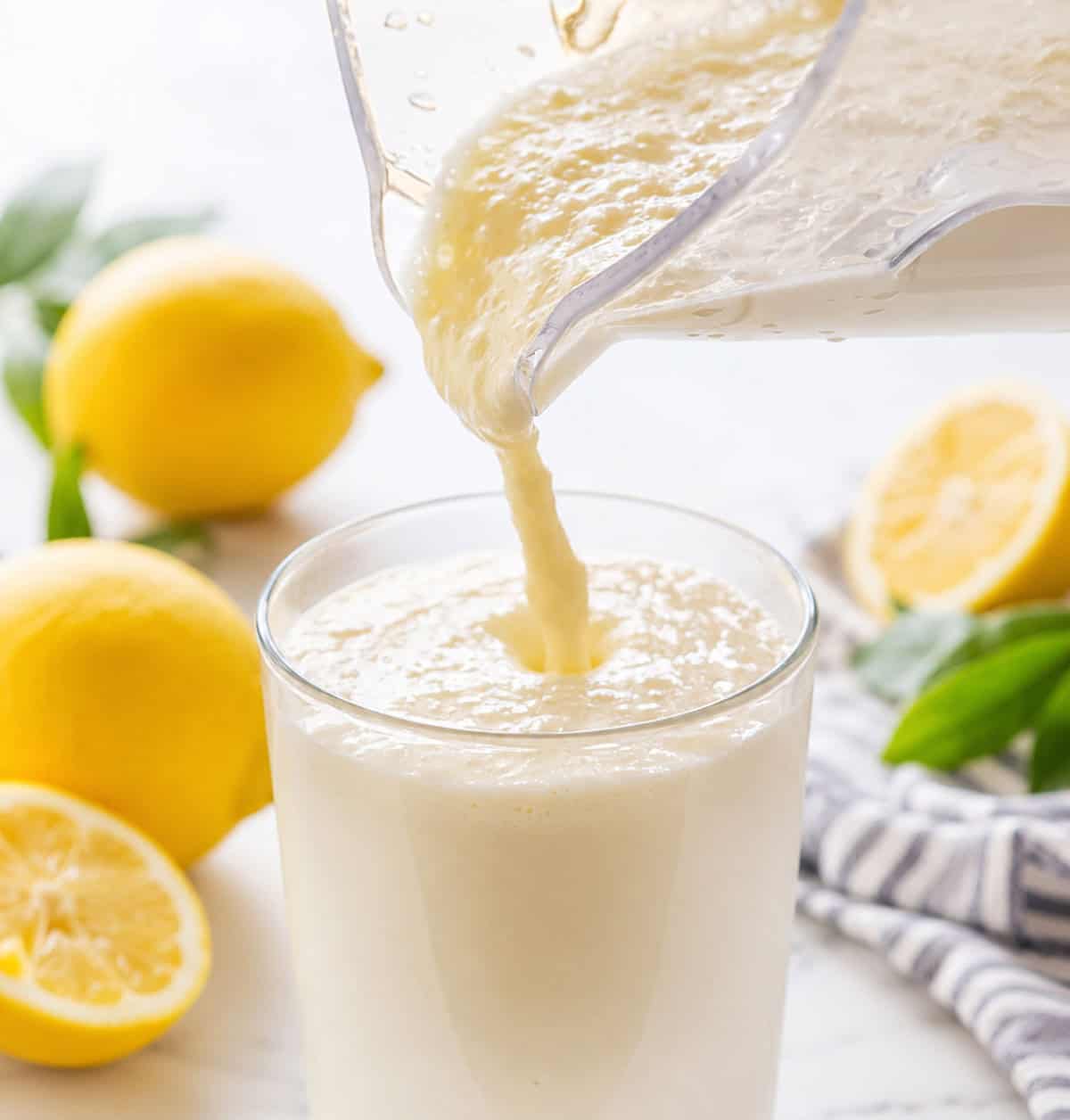 Pouring Frosted Lemonade into a glass on a white table surrounded by lemons.