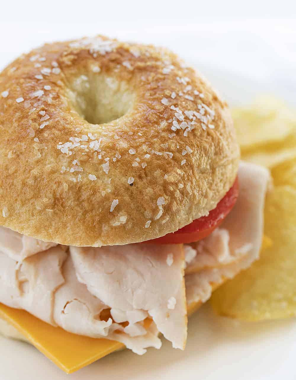 Salt Bagel Sandwich with turkey and tomato next to Chips on white plate