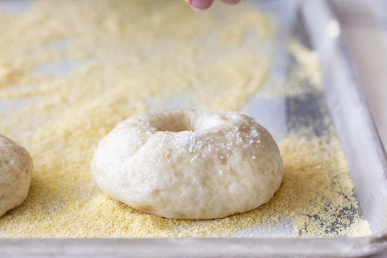 Hand Sprinkling Salt onto a Boiled Bagel and on a sheet pan with cornmeal