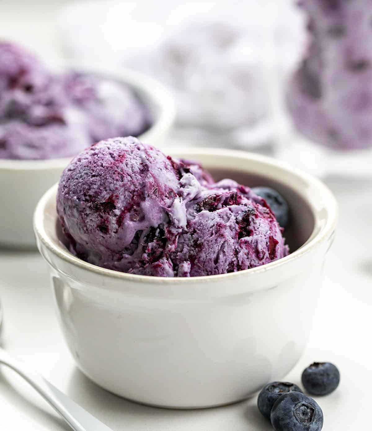 White Bowl Filled with Blueberry No-Churn Ice Cream with White Spoon and Blueberries on Counter.