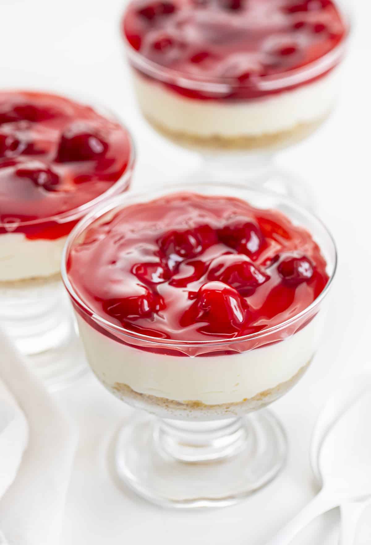 3 No-Bake Cherry Cheesecake Parfaits on a White Counter with White Spoons.