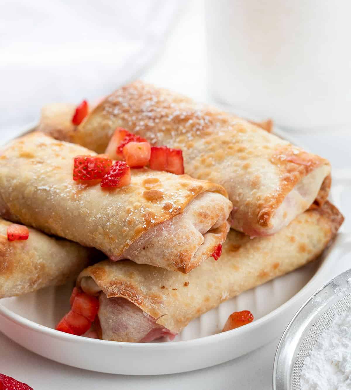 Strawberry Cheesecake Egg Rolls Stacked on a Plate with Strawberry Pieces on Top for Garnish