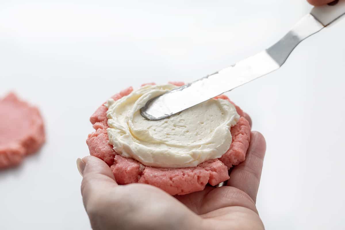 Smoothing Swiss Meringue Frosting Over a Pink Velvet Cookie.