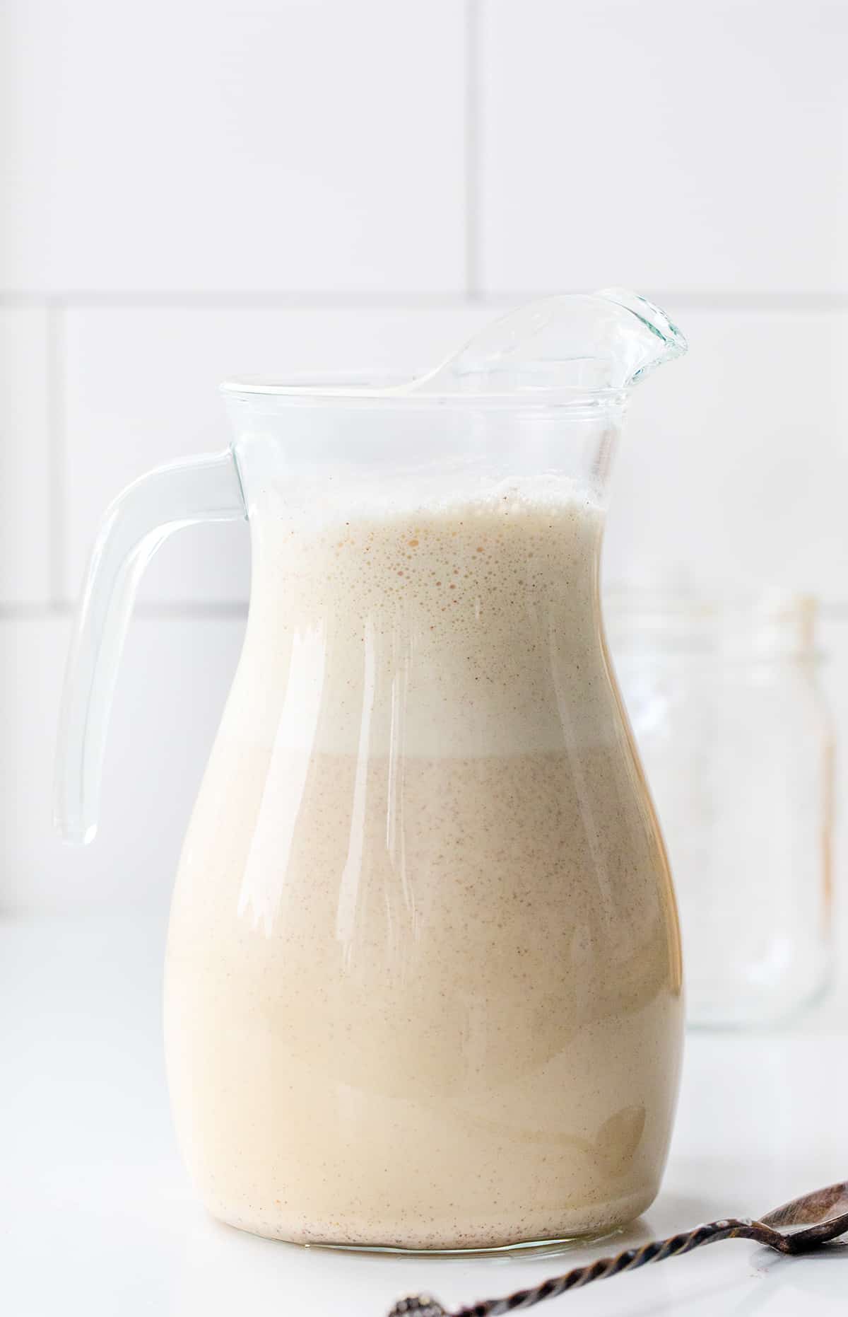 Pitcher of Homemade Rum Chata with froth neat the top of the glass pitcher on a white counter. 