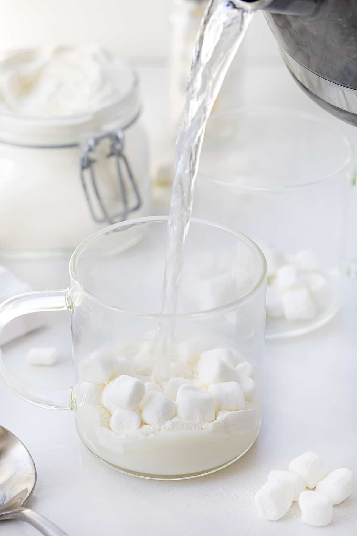 Pouring Hot water over white chocolate hot cocoa mix and mini marshmallows in a clear glass.