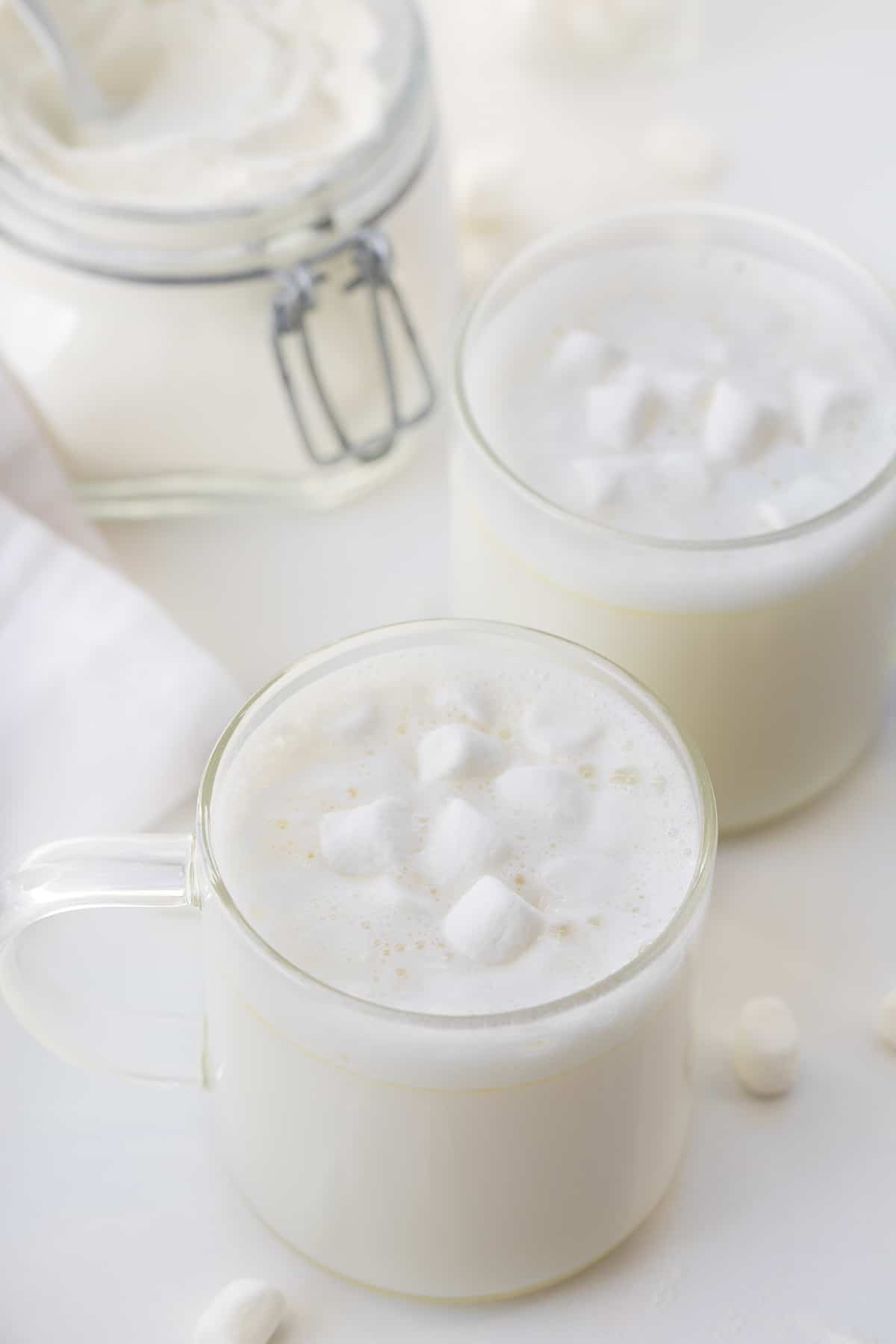 Cups of white hot chocolate on a white counter with mini marshmallows and the cocoa in the background.