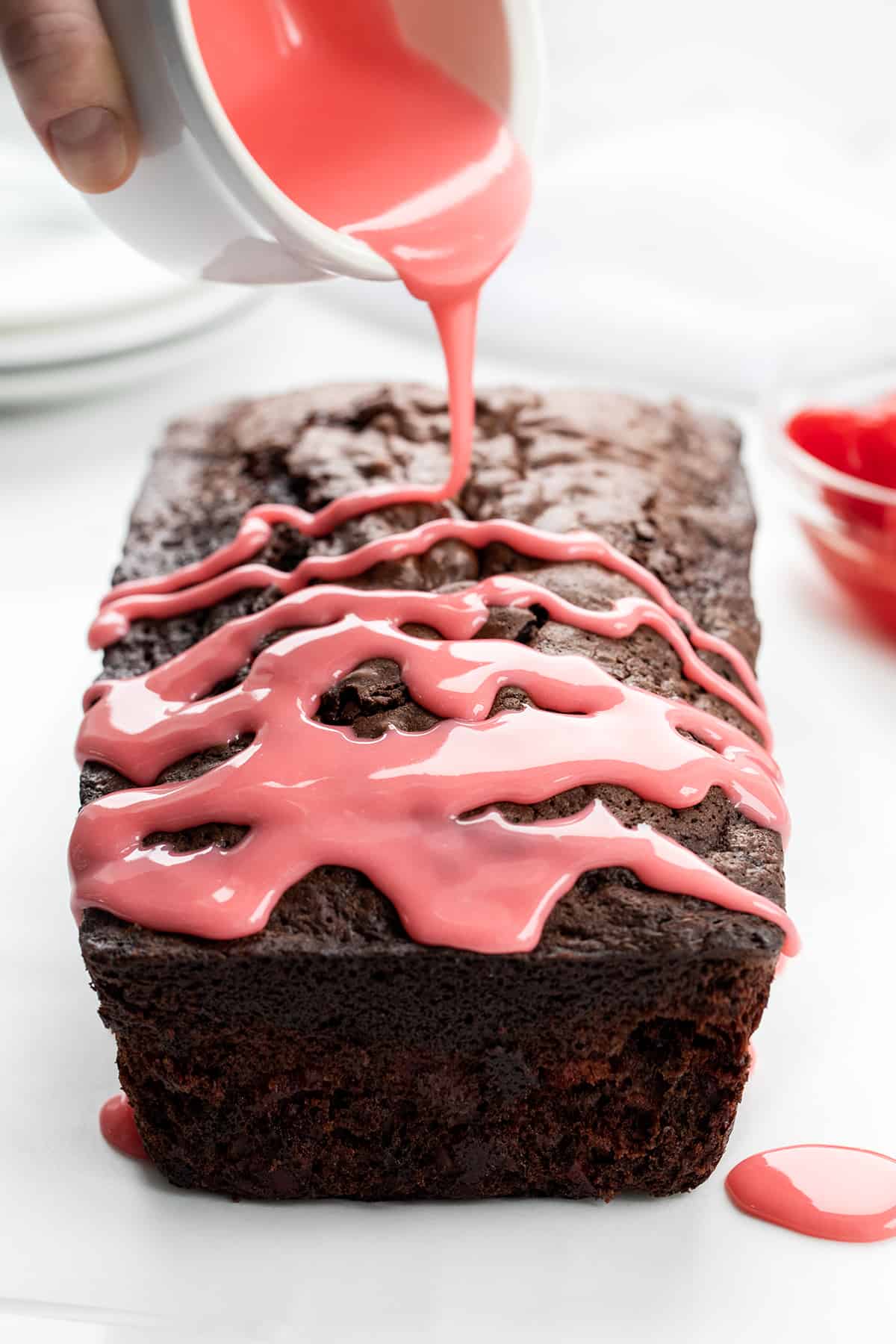Drizzling Cherry Glaze Over Cherry Brownie Bread on a White Counter.