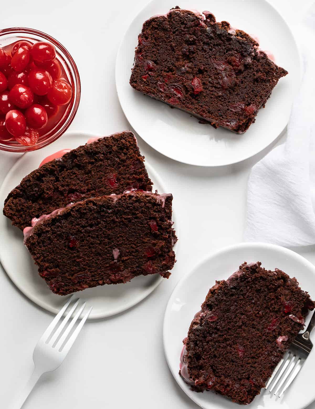 Slices of Cherry Brownie Bread on Plates on a White Counter with a Small Bowl of Cherries.