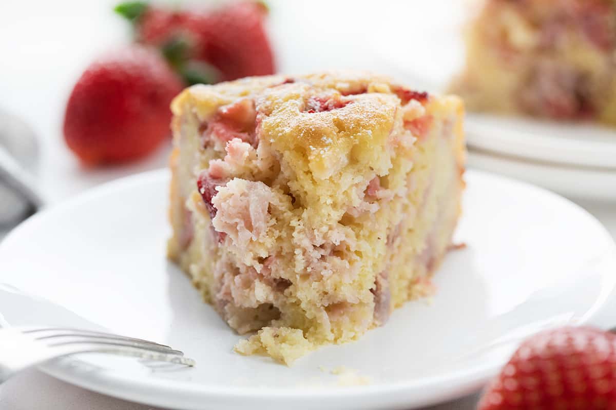 Piece of Fresh Strawberry Cake on a White Plate with Strawberries in the Background.