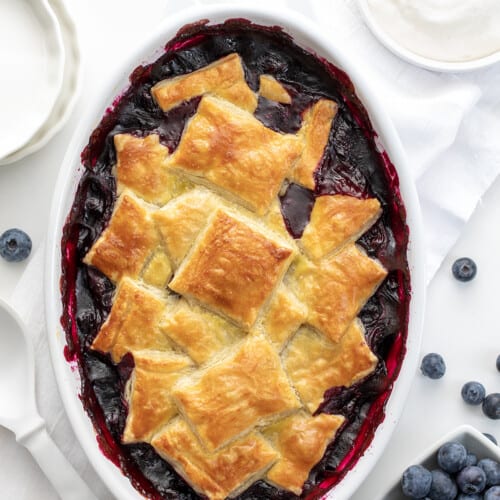 Pan of Blueberry Puff Pastry Bake from Overhead Surrounded by Blueberries and white Dishes.