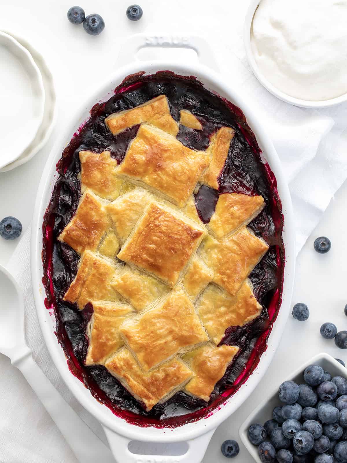 Pan of Blueberry Puff Pastry Bake from Overhead Surrounded by Blueberries and white Dishes.
