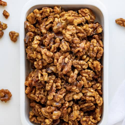 Candied Walnuts in a White Dish with Some Scattered Around and a White Towel Tucked at the bottom.