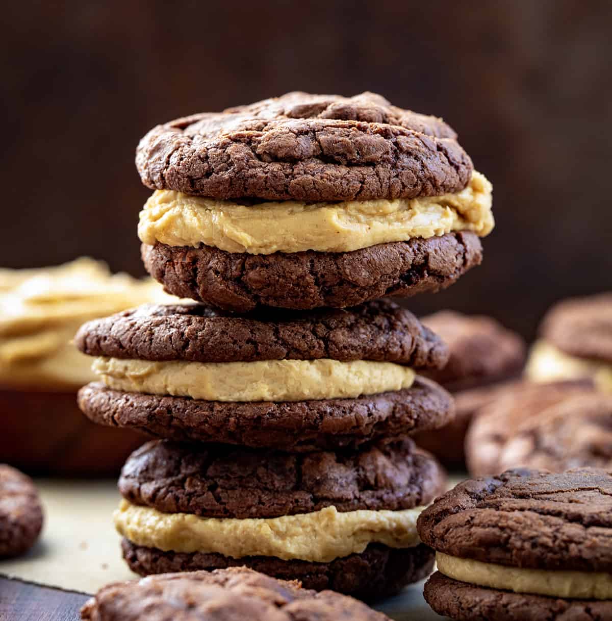 Stacked Chocolate Peanut Butter Sandwich Cookies on a Dark Cutting Board Surrounded by More Cookie Sandwiches.