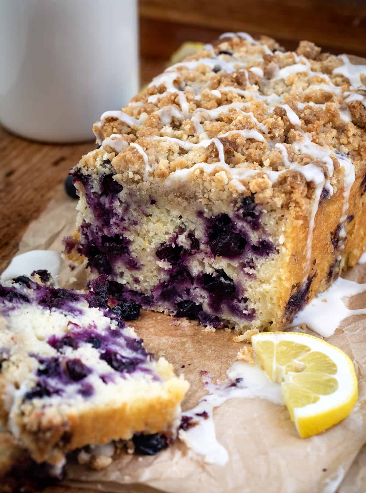 Cut Into Blueberry Lemon Loaf with Glaze and Lemons on Parchment on a Cutting Board.