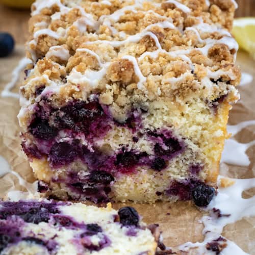 Cut Into Blueberry Lemon Loaf with Glaze and Lemons on Parchment on a Cutting Board.