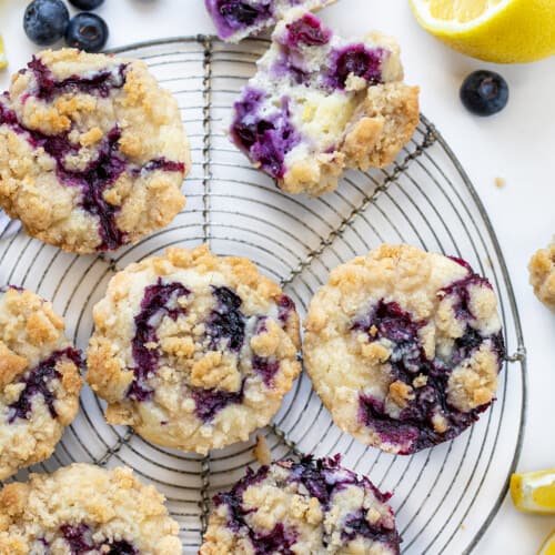 Blueberry Lemon Crumb Muffins on a Circular Wire Rack Next to a Halved Muffin with Fresh Lemon and Blueberries.