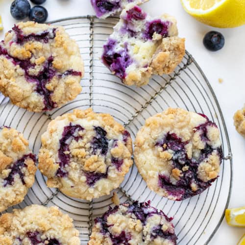 Blueberry Lemon Crumb Muffins on a Circular Wire Rack Next to a Halved Muffin with Fresh Lemon and Blueberries.