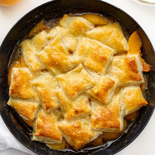 Peach Puff Pastry Bake in a Skillet on a White Counter with Fresh Peaches Around.