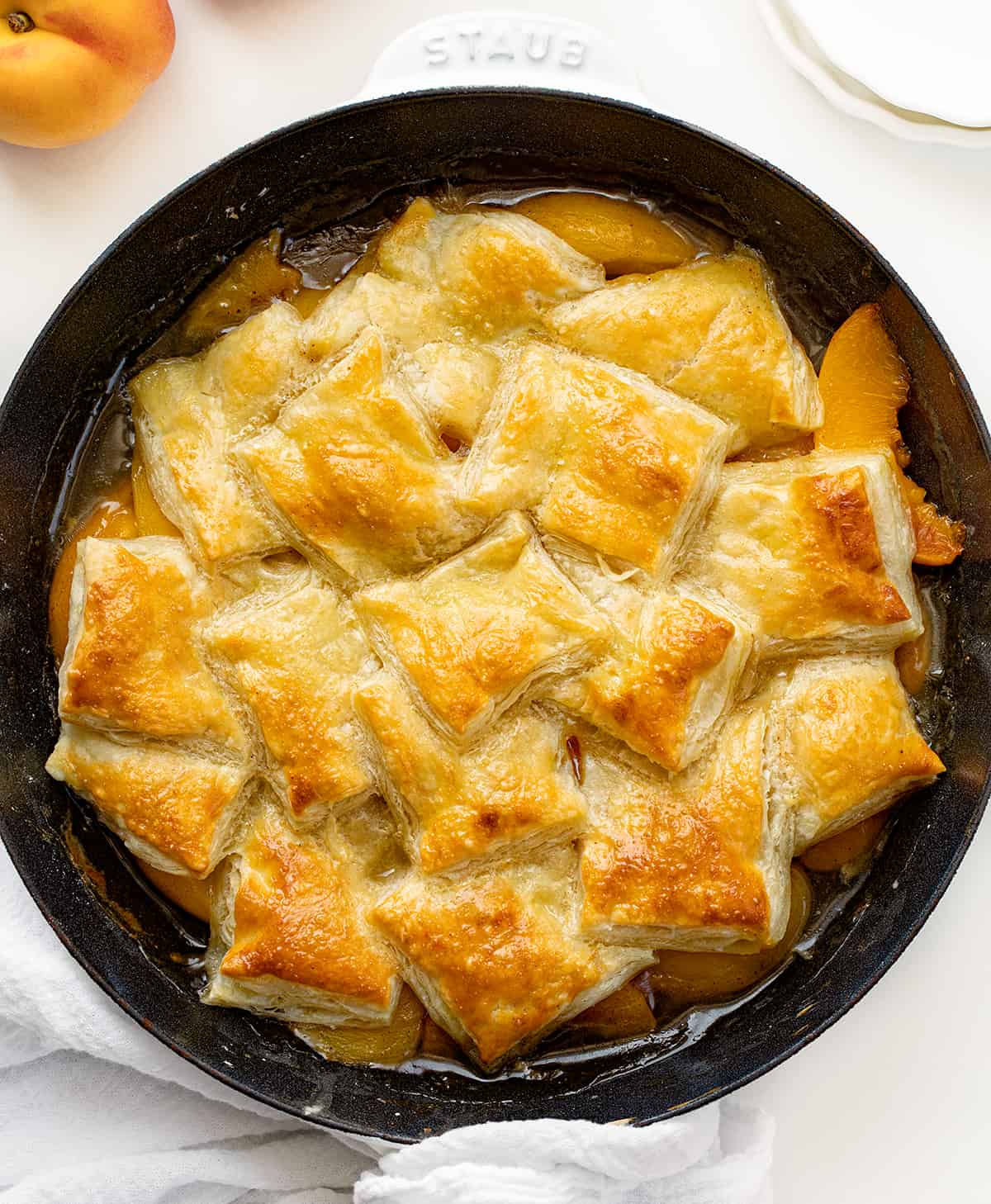 Peach Puff Pastry Bake in a Skillet on a White Counter with Fresh Peaches Around.