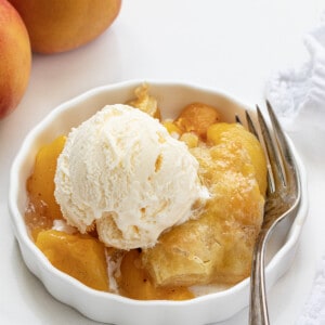 One Bowl of Peach Puff Pastry Bake on a White Counter with Peaches and a Fork and a Scoop of Vanilla Ice Cream.