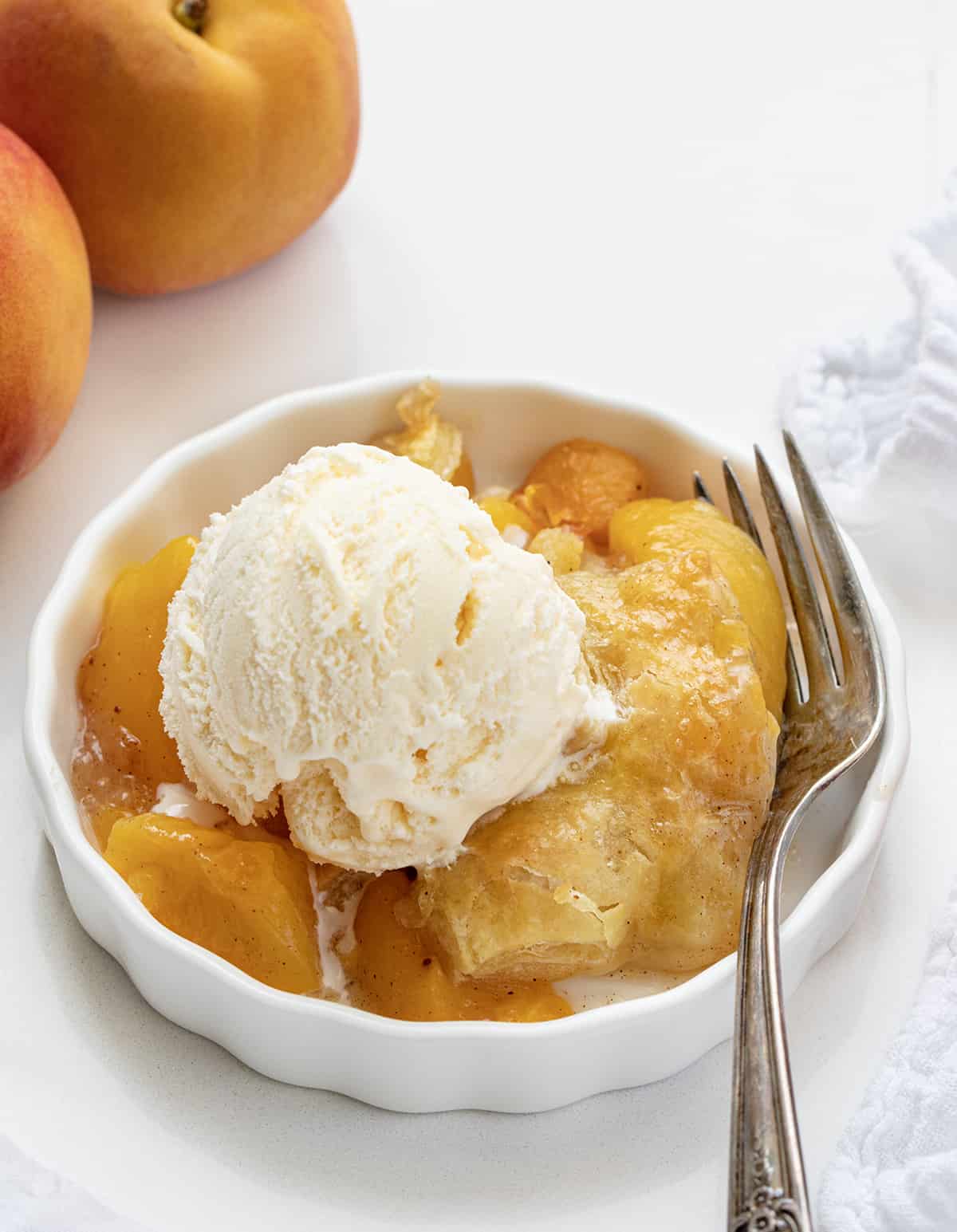 One Bowl of Peach Puff Pastry Bake on a White Counter with Peaches and a Fork and a Scoop of Vanilla Ice Cream.