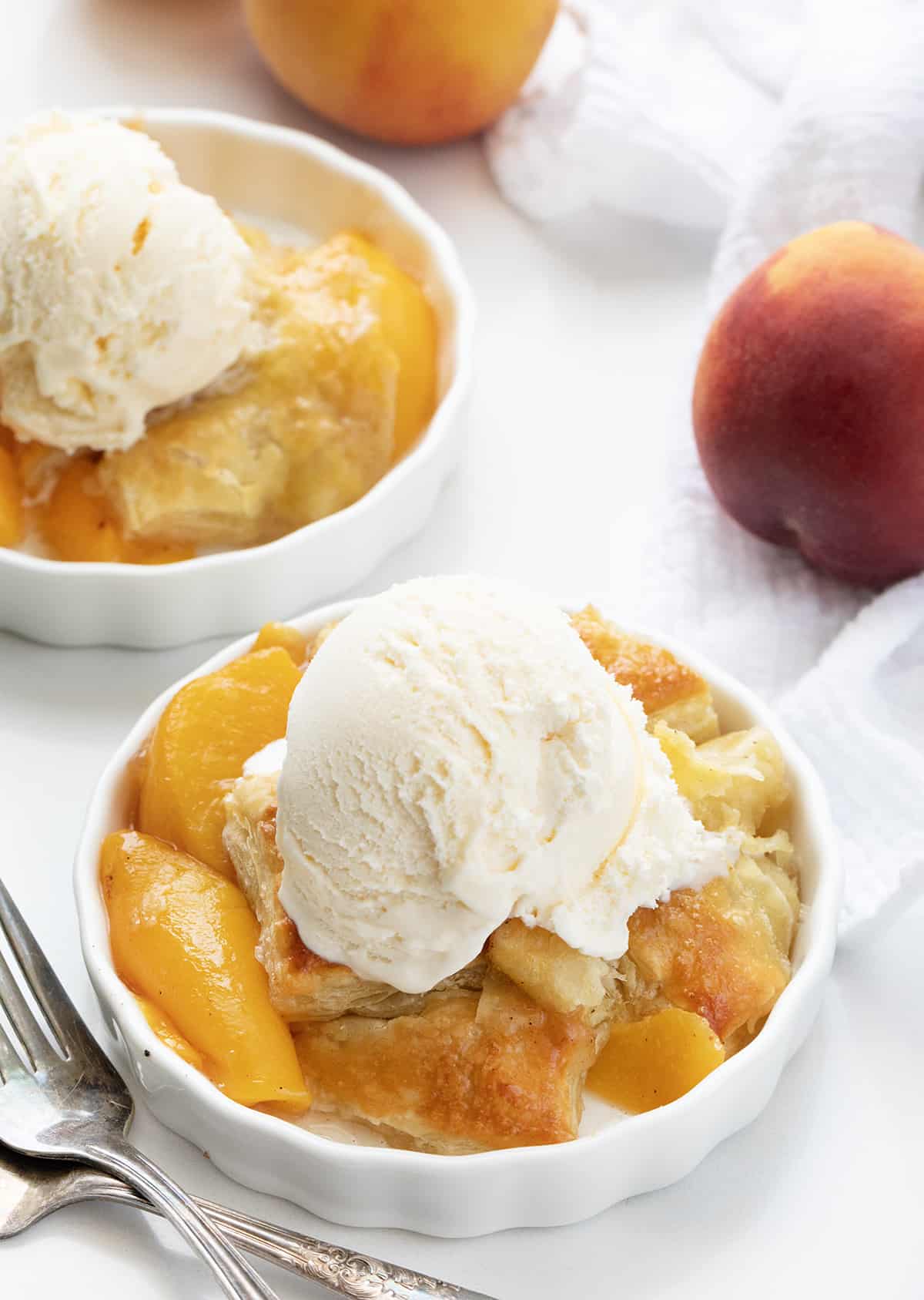 Two Bowls of Peach Puff Pastry Bake on a White Counter with a Fresh Peach and Vanilla Ice Cream Scooped on Top.